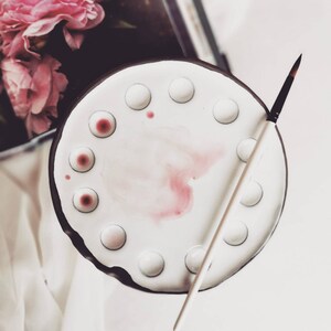 Op de afbeelding: Een wit keramiek verfpalette met 12 vakjes, waarvan sommige gevuld zijn met roze verf. Een wit penseel ligt op het palette.