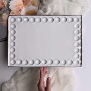 May include: A rectangular white ceramic serving tray with a brown rim and a border of raised, circular accents. The tray is held by a hand, with a floral print and sheer fabric in the background.