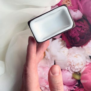 以下が含まれることがあります： 濃い茶色の縁取りのある小さな白い陶器の皿。皿はピンクと白の花の背景に手の中で持たれている。