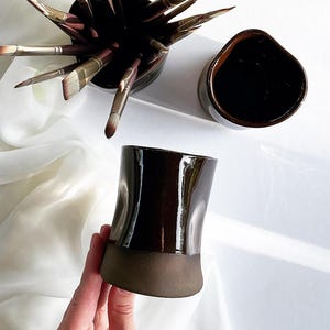Peut inclure: Un ensemble de pinceaux dans un pot en céramique marron foncé, avec une tasse et un vase en céramique marron foncé assortis. Le vase est tenu par une main. Les objets sont sur une surface blanche.