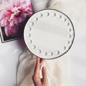 May include: White ceramic plate with a brown rim and a circle of small indentations around the edge. The plate is sitting on a white fabric surface.