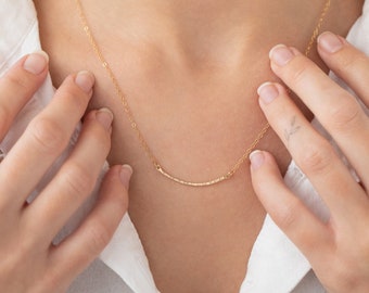 May include: A delicate gold necklace with a thin, curved bar pendant. The necklace is worn by a person with their hands near their neck.