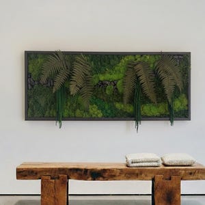 May include: A rectangular framed wall art piece featuring a moss and fern arrangement. The artwork has a dark frame and includes various shades of green moss and faux ferns. A wooden bench with two folded cream-colored blankets sits below the artwork.