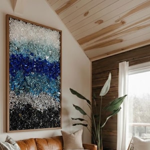 May include: A large rectangular wall art piece featuring a layered mosaic of crushed glass in shades of white, light blue, deep blue, silver, and black, encased in a simple wooden frame, displayed vertically on a plain wall above a leather sofa.