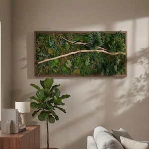 May include: A rectangular wall art piece featuring a dense arrangement of preserved green moss, various small ferns, and a light-colored wooden branch set within a thin, dark brown wooden frame, mounted horizontally on a neutral-toned wall.