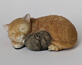 May include: A ceramic figurine of a sleeping orange tabby cat curled up with a smaller gray kitten. The cat's eyes are closed and its paws are tucked under its body.