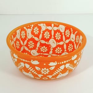 May include: Orange ceramic bowl with a white floral and fishbone pattern. The bowl has a rounded shape and a smooth, glossy finish. The interior and exterior are decorated with the repeating design. A decorative piece for the kitchen or dining room.