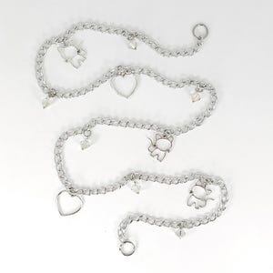 May include: Silver-colored chain accessories with heart and cat charms, and clear crystal accents. The chains are arranged in a wavy pattern against a white background. The charms are small and delicate, adding a touch of whimsy.