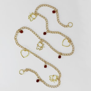 May include: Gold-colored chain belts with cat and heart charms, and small red accents. The chains are arranged on a white background, showcasing the delicate design and details of the jewelry.