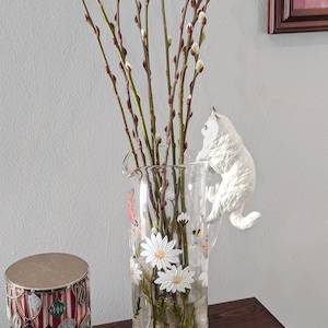 May include: A clear glass pitcher with a floral design featuring white daisies and a bee. The pitcher is filled with pussy willow branches and a white ceramic cat figurine is perched on the rim.