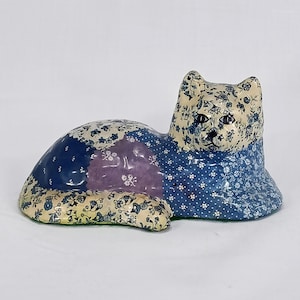 May include: A ceramic cat figurine in a reclining pose. The cat's body is a patchwork of blue, purple, and cream floral patterns. The head and front paws are detailed with a blue floral design, and the eyes are dark.
