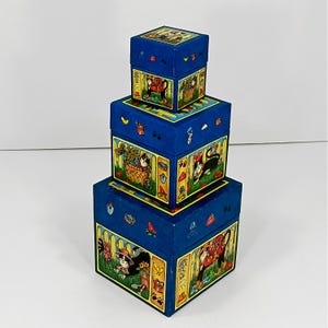 May include: A set of three blue nesting boxes with colorful illustrations of cats, flowers, and other garden elements. The boxes are stacked, with the largest at the bottom and the smallest on top. The boxes have a vintage aesthetic.