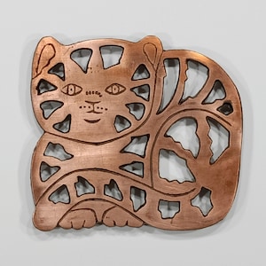 May include: A copper-colored metal cat-shaped cutout. The cat has a smiling face, eyes, and spots. The design includes cut-out shapes within the cat's body and tail. The metal has a slightly aged appearance.
