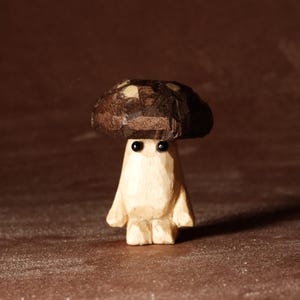 May include: A carved wooden mushroom figure with a brown cap and a light-colored body. The mushroom has two small black eyes and a whimsical, cartoonish appearance. The figure is standing on a brown surface.