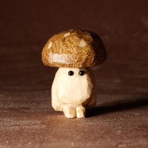 May include: A carved wooden mushroom figure with a light brown cap speckled with white dots. The stem is a light beige color, and it has two small black eyes. The figure is standing on a brown surface.