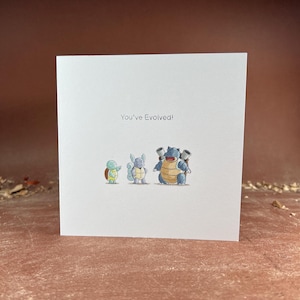 May include: A square white greeting card featuring a small illustration of three blue turtle-like creatures in different stages of growth. Above the characters, the text reads "You've Evolved!" in a simple, dark, sans-serif font.