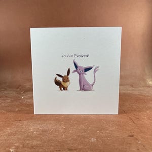 May include: A square white greeting card featuring a brown Eevee and a purple Espeon illustration. The text "You've Evolved!" is printed in black above the two characters. The card has a clean, simple design with ample white space surrounding the art.