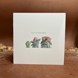 May include: A white greeting card with the text "You've Evolved!" above a watercolor illustration of three stages of a plant-like creature. The creatures are green with pink and red accents.