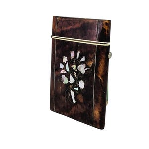 May include: A rectangular, dark brown box with a decorative floral design made of white and iridescent shell pieces. The box has a gold-colored metal band around the top and a small metal clasp on the side.