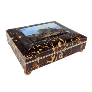 May include: A rectangular tortoiseshell box with a hinged lid. The lid features a painted scene of a building with a dome. The box has a small keyhole and rests on four wooden feet.