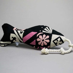 May include: Black and pink patterned fish-shaped toy with a white rope tail. The toy is made of fabric and has a geometric design.
