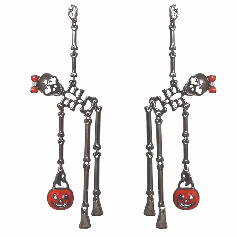 May include: A pair of Halloween-themed earrings. Each earring features a skeleton design with a skull, rib cage, and dangling bone accents. A small red bow adorns each skull. A smiling orange pumpkin charm hangs from each earring.