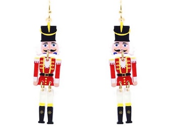 Puede incluir: Un par de pendientes colgantes festivos con un diseño de soldado cascanueces. Cada pendiente presenta una chaqueta roja, un sombrero negro y cabello blanco, con detalles dorados. Los pendientes están suspendidos de ganchos dorados.