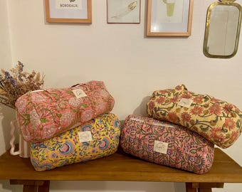 Peut inclure: Plusieurs sacs de voyage à motifs floraux, dans des tons de rose, jaune et crème, sont disposés sur une surface en bois. Les sacs présentent un design matelassé et une petite étiquette. Des œuvres encadrées et un miroir décoratif sont en arrière-plan.