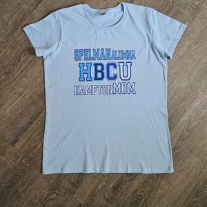 May include: A light blue t-shirt with the text "SPELMAN ALUMNA HBCU HAMPTON MOM" in shades of blue. The shirt is made of a soft material and has short sleeves. The text is centered on the front of the shirt.