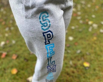 Könnte beinhalten: Graue Jogginghose mit dem Wort "SPELMAN" vertikal auf dem linken Bein in einem Blau- und Türkis-Farbverlauf gedruckt.