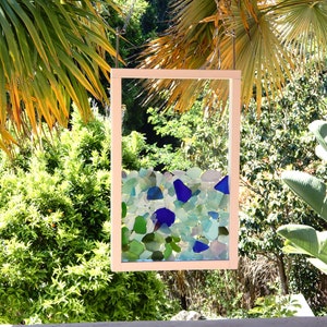 May include: A white framed window with a mosaic of blue and green sea glass inside. The window is hanging from a chain and is surrounded by green foliage.