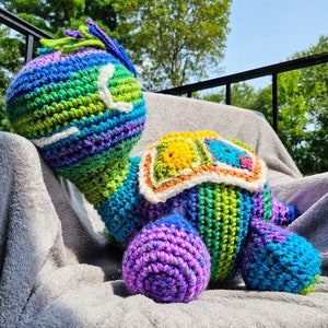 May include: A large, handmade crochet turtle in vibrant rainbow colors. The turtle features a detailed shell, head, and limbs, with a textured, yarn-based design. The turtle is resting on a gray blanket, with a natural outdoor background.