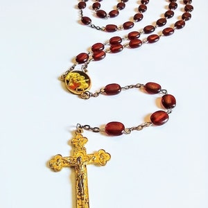 May include: A rosary with oval, reddish-brown beads strung on a silver-colored chain. The rosary features a gold-colored crucifix and a gold-colored medallion with an image. The crucifix has a detailed depiction of Jesus.