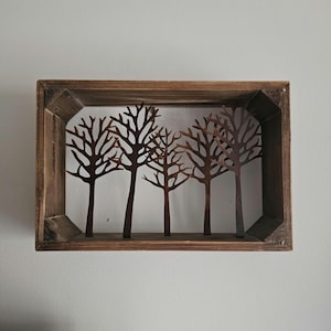 May include: A rustic wooden shadow box featuring five stylized metal tree silhouettes. The trees are a dark brown color, set against a white background, and framed by a weathered wooden box. The box is mounted on a light gray wall.