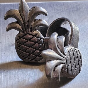 May include: Two silver-colored pineapple-shaped napkin rings. The rings have a textured surface and are designed with detailed leaves and a diamond pattern on the pineapple body. The rings are on a silver surface.