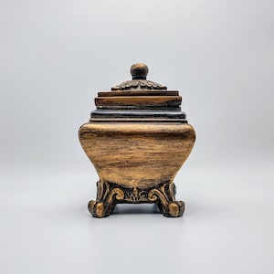 May include: A decorative, lidded box with a vintage aesthetic. The box is primarily gold with dark brown accents and rests on ornate, scrolled feet. The lid features a small, round finial and decorative trim. The box has a classic, antique look.