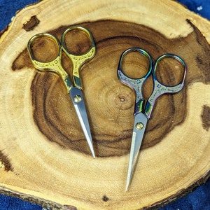 May include: Two pairs of small scissors on a wooden surface. One pair is gold with a floral design, the other is iridescent with a floral design.