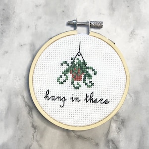 May include: A cross-stitch embroidery hoop featuring a green and red plant in a pot, with the words "hang in there" stitched below. The embroidery is set in a wooden hoop, perfect for home decor.