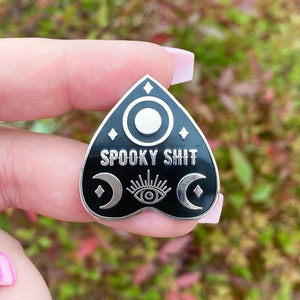 May include: Black and silver enamel pin in a planchette shape. The pin features the text "SPOOKY SHIT" and crescent moons, an eye, and a circle design. The pin is held in a hand.