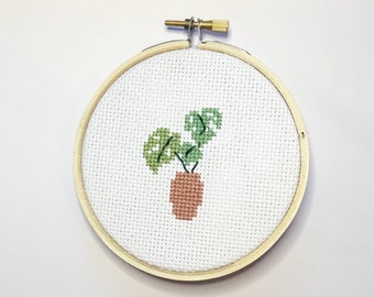 May include: A cross-stitch embroidery of a potted plant with green leaves and a brown pot. The embroidery is set in a round wooden hoop, perfect for wall decor. The background is white.
