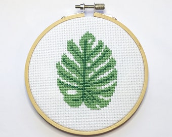 May include: A cross-stitch embroidery of a green monstera leaf, set in a round wooden hoop. The leaf design is centered on a white fabric background. The hoop has a metal tightening mechanism.