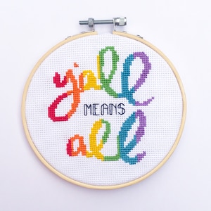 Op de afbeelding: Een borduurring met de tekst "y'all means all" in regenboogkleuren. De tekst is geborduurd op witte stof en wordt omlijst door een houten borduurring. De tekst is in een cursief lettertype.