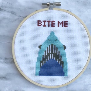 May include: A cross-stitch embroidery hoop featuring a shark design with the words "BITE ME" in red. The shark is depicted with shades of blue and gray, set against a white background. The wooden hoop is round.