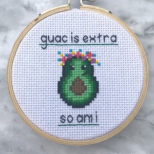 May include: Cross-stitch embroidery in a wooden hoop features a green avocado with a floral crown. The text reads "guac is extra so am I." The design is on white fabric with a cross-stitch pattern. The artist's name is at the bottom.