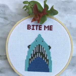 May include: A cross-stitch hoop featuring a shark's open mouth with the words "BITE ME" in maroon. The shark is in shades of blue and green, set against a white background. A small succulent plant is attached to the top of the hoop.