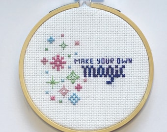 May include: A cross-stitch embroidery hoop with the words "MAKE YOUR OWN magic" stitched in blue thread. The design includes colorful starbursts in pink, green, and blue. The hoop is made of wood and is approximately 6 inches in diameter.