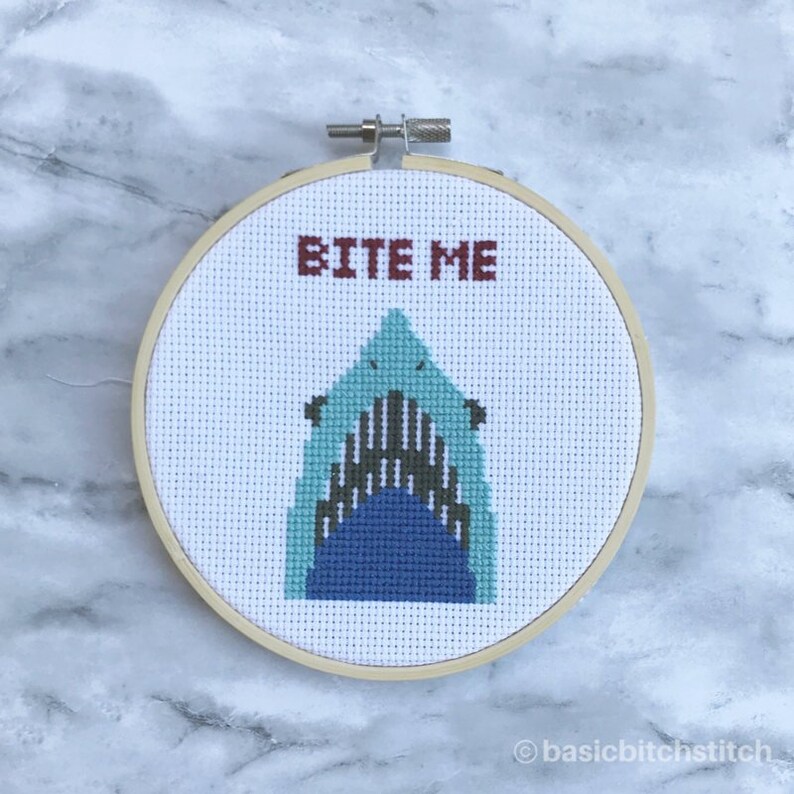 May include: A cross-stitch embroidery hoop featuring a shark design with the words "BITE ME" in red. The shark is depicted with shades of blue and gray, set against a white background. The wooden hoop is round.