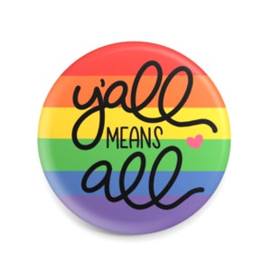 May include: A round button with a rainbow background and the words "y'all MEANS all" in black script. A small pink heart is next to the word "all". The button is a symbol of inclusivity and support for the LGBTQ+ community.