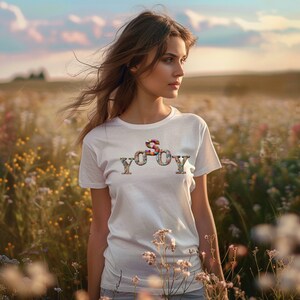 Puede incluir: Una camiseta blanca con un diseño gráfico colorido que dice "SOY YOY". La camiseta la lleva puesta una persona de pie en un campo de flores.