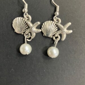May include: A pair of silver-toned earrings featuring seashell and starfish charms, with a dangling white pearl. The earrings have a hook closure. The jewelry is designed with a beach or nautical theme.